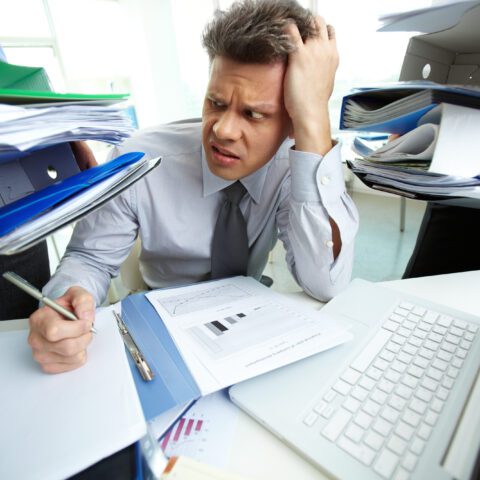 Frustrated office worker sitting at a desk covered in stacks of paper, struggling to locate a file.